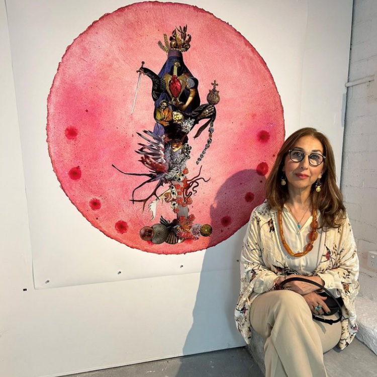 Photo of Shahroud sitting in front of one of her artworks, she wears light tan coloured warm toned clothing, round glasses, long hair, beads and drop earrings. Her artowrk is a round pink water coloured looking circle that has a  decorative and ecclectic arrnagement running down the centre, there are objects like swords, birds, bones, shells and red gemstones 